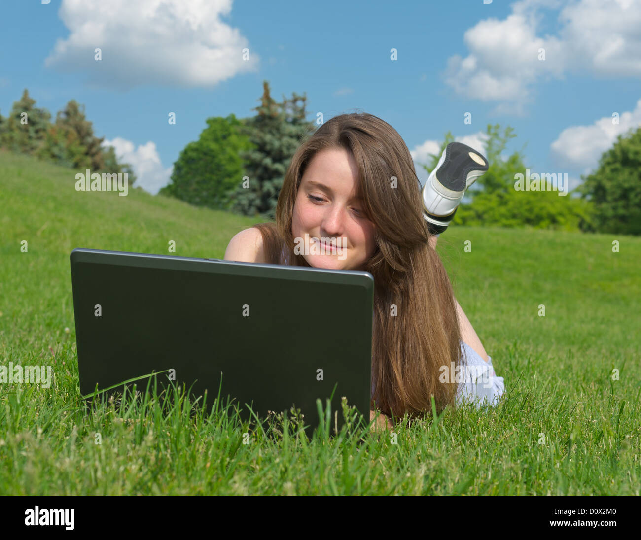 Smiling young woman lying on her stomach in the grass using a laptop outdoors Stock Photo