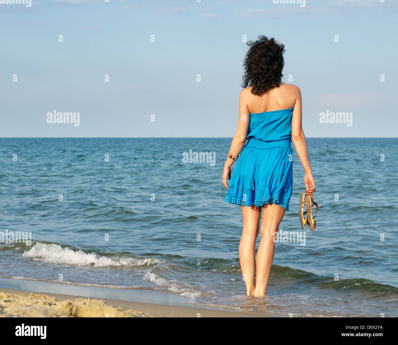 Woman in a blue summer dress standing holding her sandals in the shallow surf looking over ocean Stock Photo