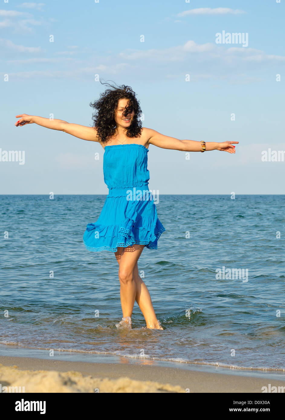 Woman in a blue summer dress dancing in the shallow sea with her arms outspread enjoying the freedom of a summer vacation Stock Photo