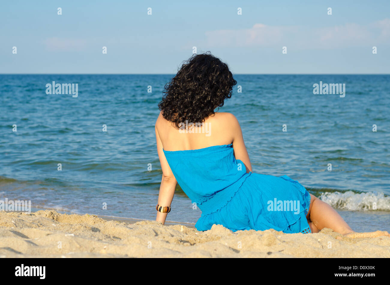 Woman with beautiful curly brunette hair sitting on a sandy beach with her back to the camera admiring the ocean Stock Photo