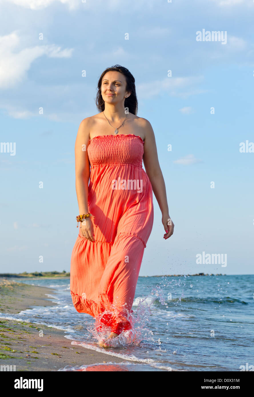 Attractive middle-aged woman in a long orange summer dress walking through surf at the edge of a beach in summer sunshine Stock Photo