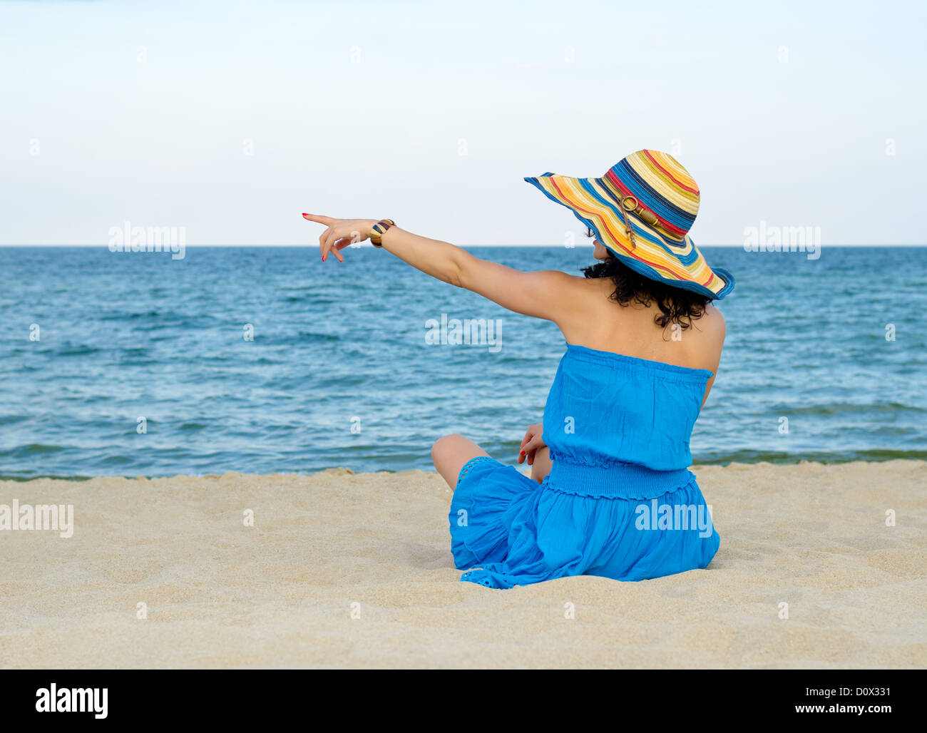 Rear view of a woman in a colourful straw sunhat sitting on a sandy beach pointing to the ocean Stock Photo
