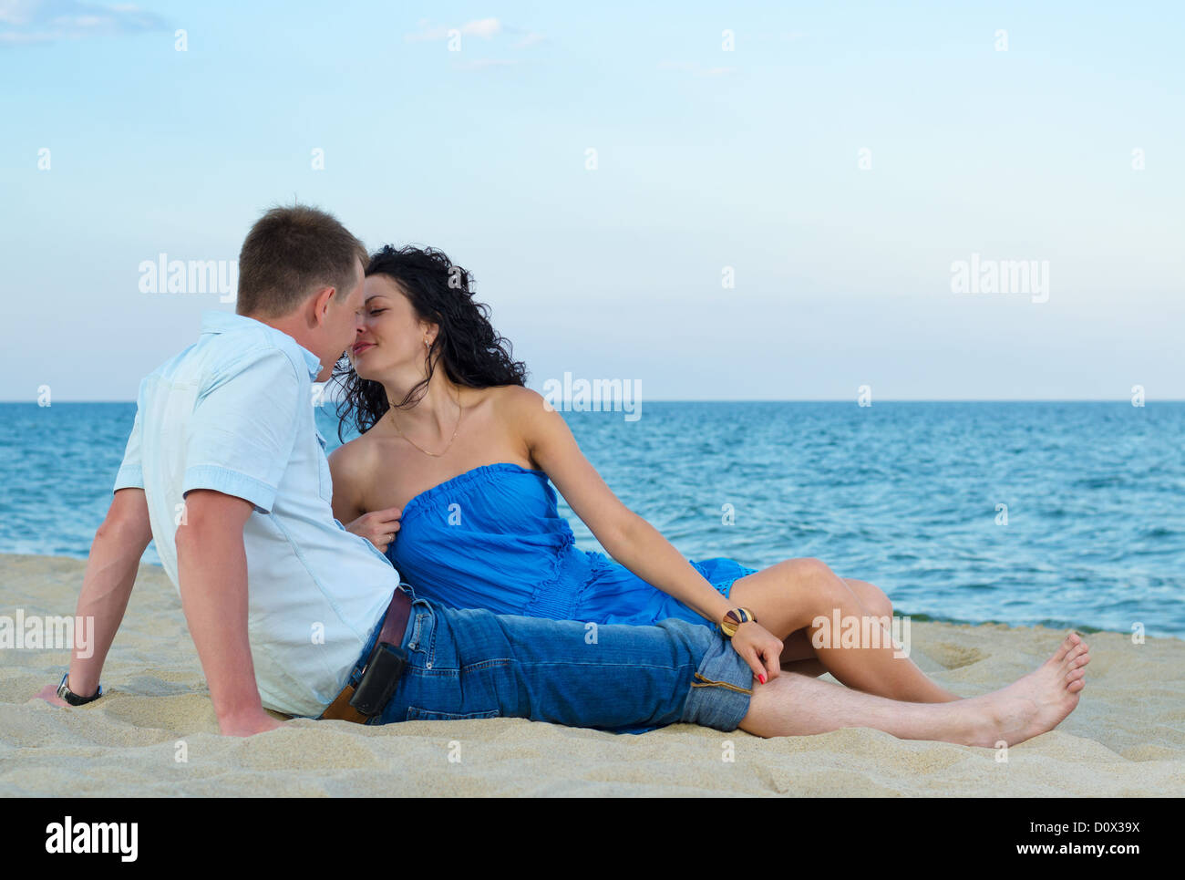 Romantic young couple sitting in the sand and kissing at the seaside with an ocean backdrop Stock Photo