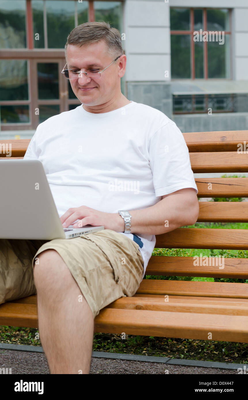 Middle-aged man sitting on wooden bench using a laptop in front of an urban building Stock Photo