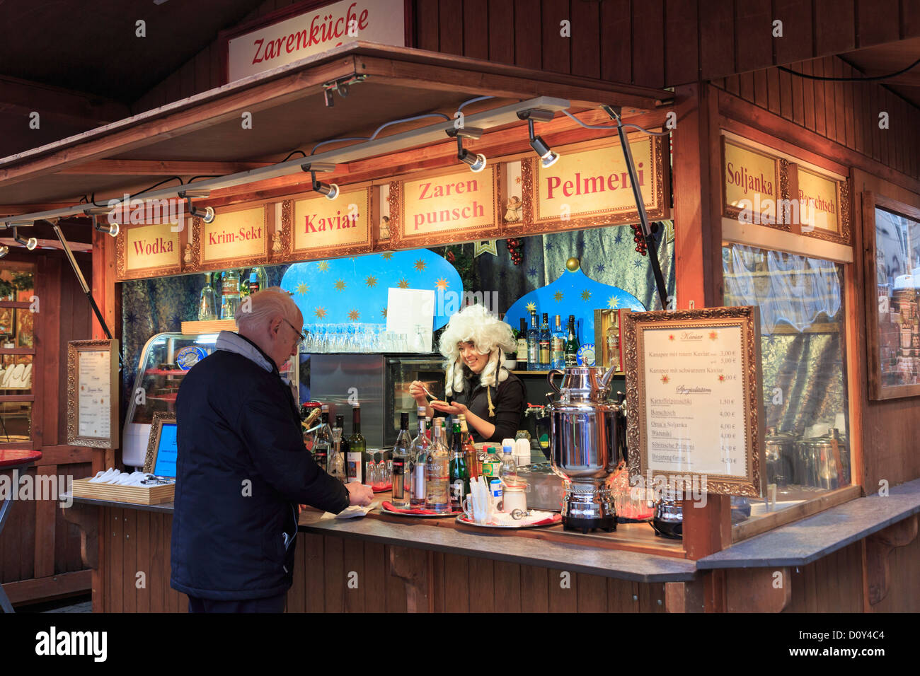 Man buying a snack from a smiling woman wearing a wig on a traditional Christmas market stall selling food and drinks Alexanderplatz, Berlin, Germany Stock Photo