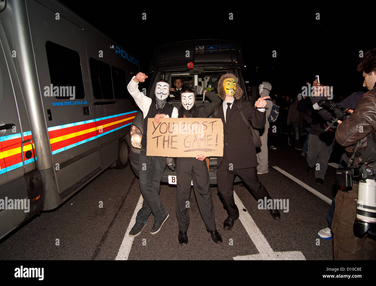 A group of anonymous masked protesters stop to stand and pose for media photographers in front of a police riot transit. Stock Photo