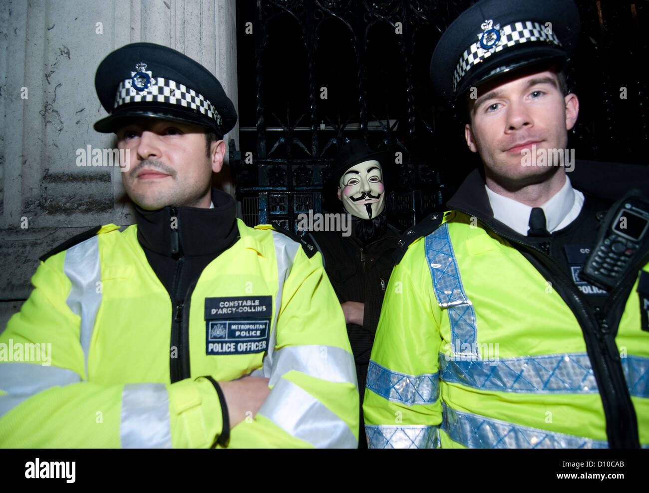 A 'Anonymous' protester stands behind police constables outside the Houses of Parliament during operation Vendetta protest Stock Photo