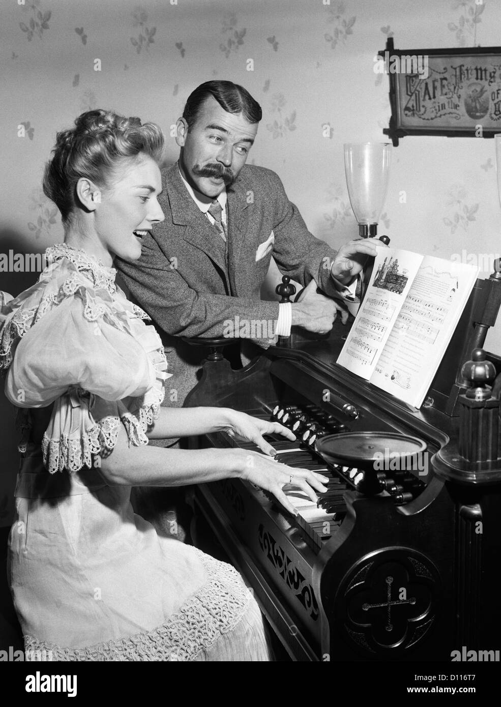 1900s COUPLE IN TURN OF THE CENTURY DRESS WOMAN PLAYING PARLOR ORGAN MAN WITH HANDLEBAR MUSTACHE TURNS PAGES OF SHEET MUSIC Stock Photo
