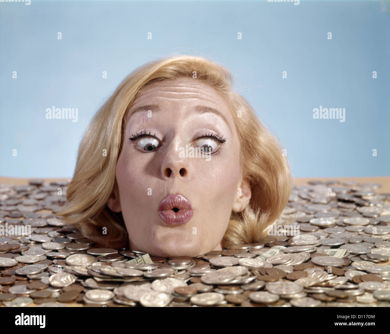 1960s BLOND WOMAN FUNNY FACIAL EXPRESSION DROWNING IN COINS UP TO HER NECK Stock Photo