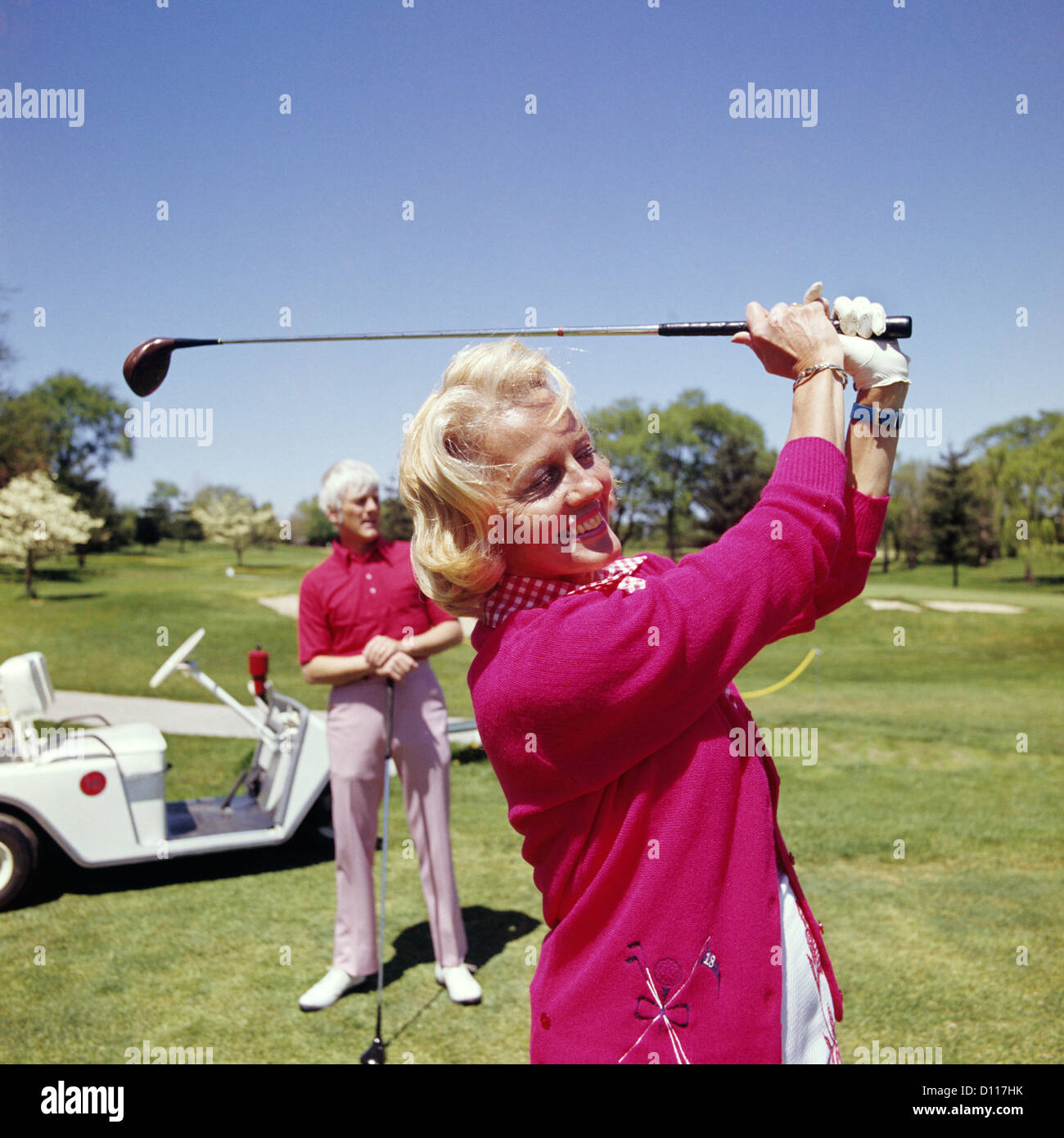 1960s MATURE WOMAN GOLFING AS MAN STANDS BY GOLF CART Stock Photo