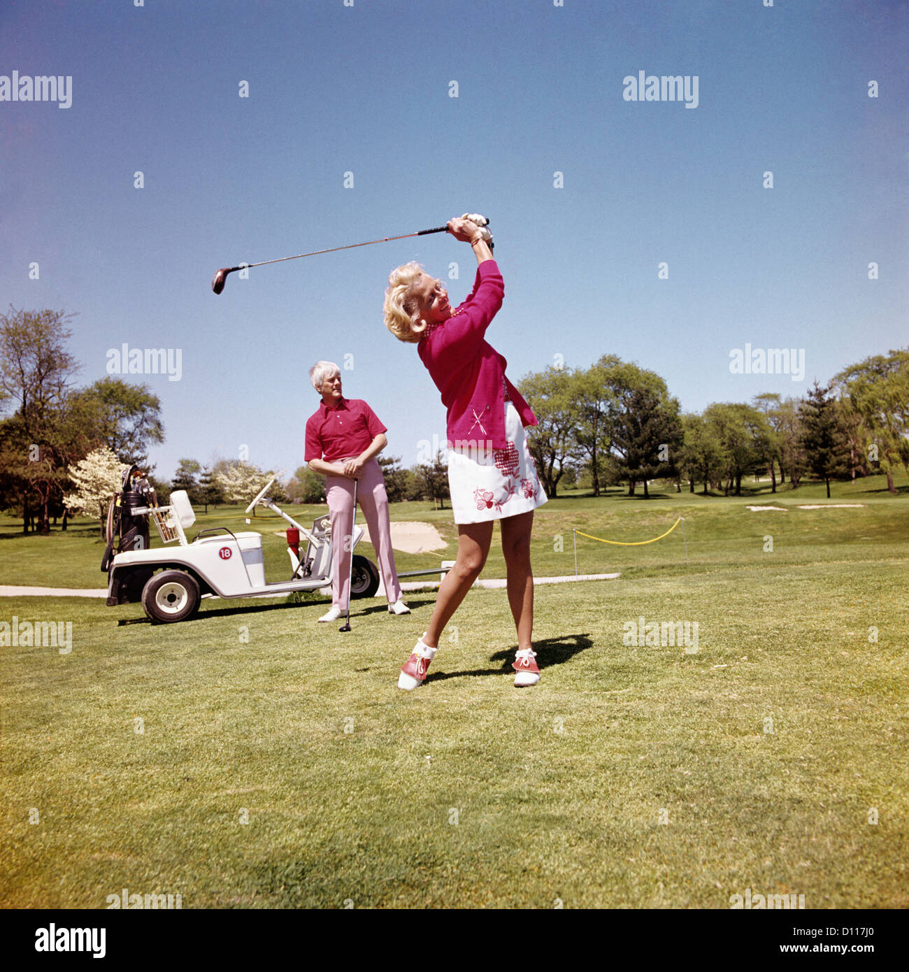 1970s MATURE WOMAN SWINGING GOLF CLUB AS MAN STANDS BY GOLF CART Stock Photo