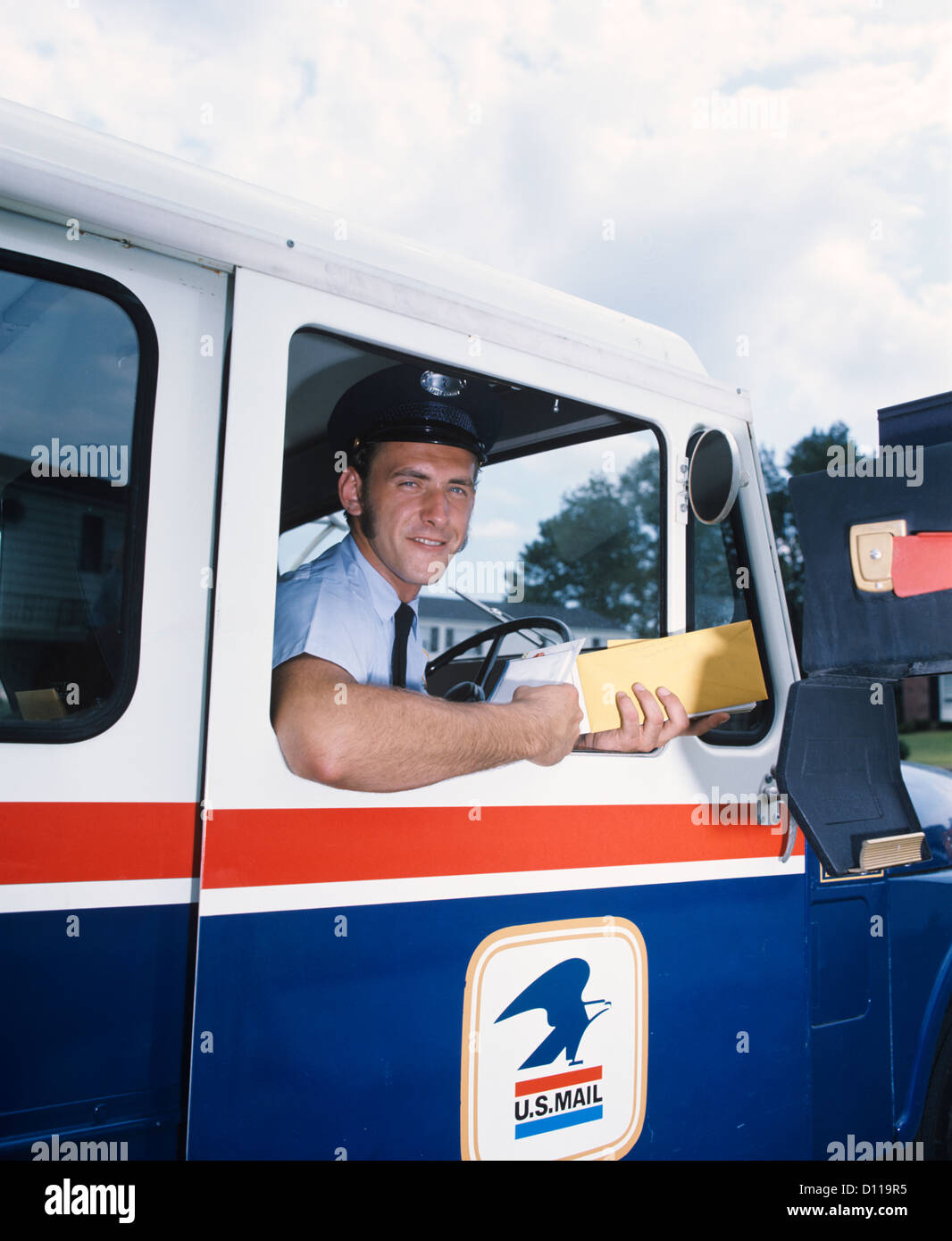 1970s SMILING MAILMAN IN U.S. MAIL TRUCK DELIVERING MAIL TO HOME MAILBOX LOOKING AT CAMERA Stock Photo
