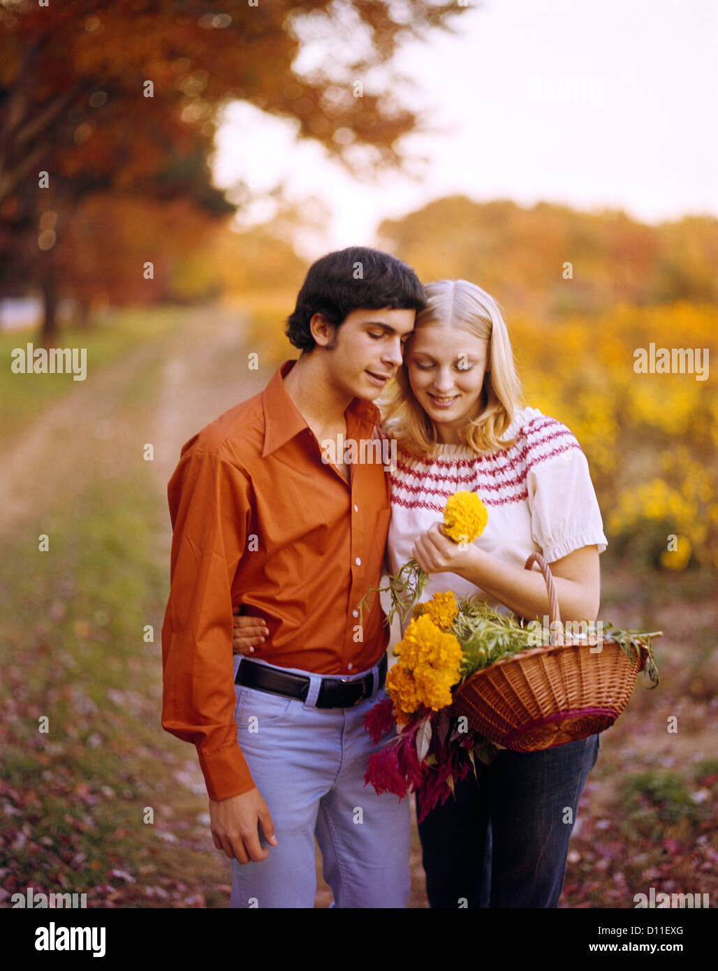 1970s AFFECTIONATE YOUNG COUPLE WITH BASKET OF FLOWERS ON AUTUMN COUNTRY ROAD Stock Photo
