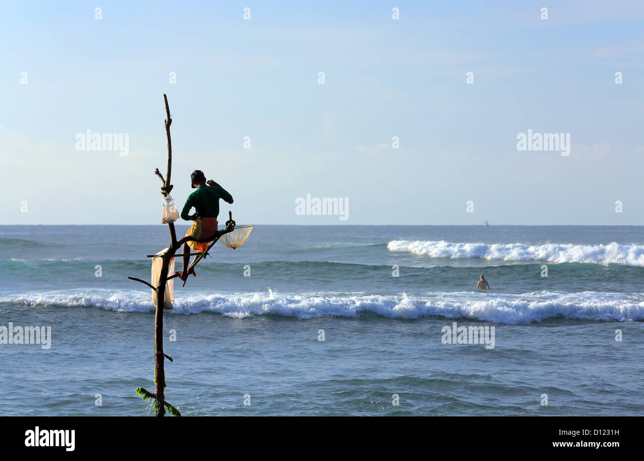 Traditional stick fisherman early morning fishing in Weligama Bay with a surfer waiting for waves in the background. Stock Photo