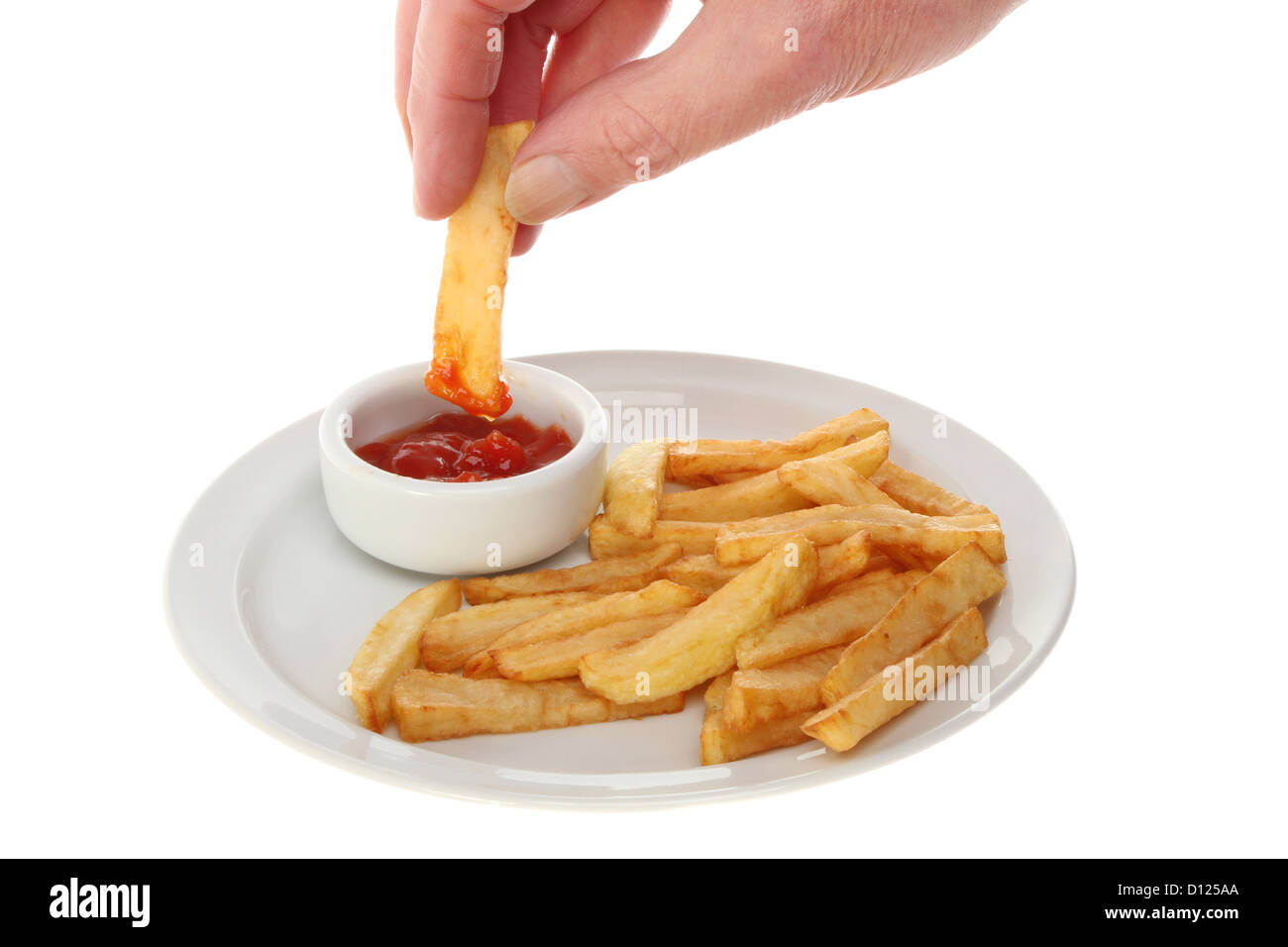 Hand dipping a French fry into tomato ketchup in a ramekin next to fries on a plate isolated against white Stock Photo