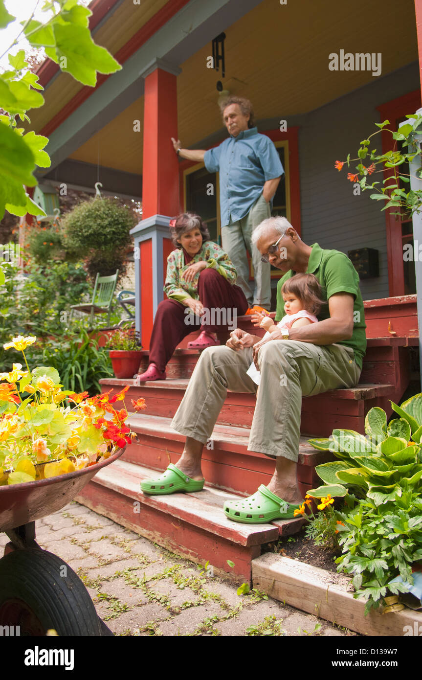 Family Sitting On Front Porch Of Home Surrounded By Plants And Flowers