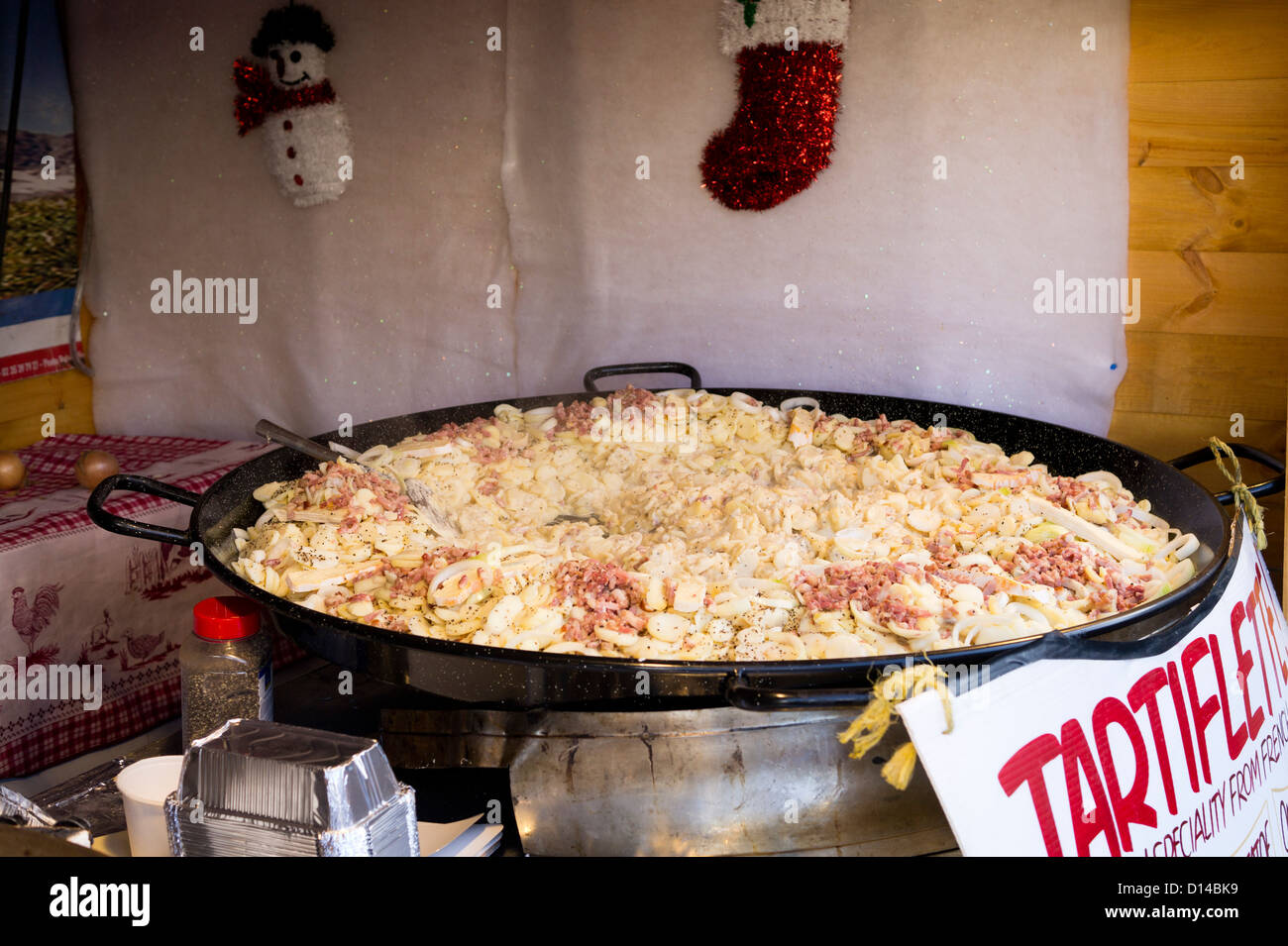 Christmas market stall cooking traditional French food Tartiflete in a giant frying pan. Stock Photo