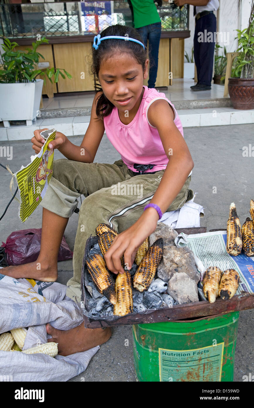 A young Filipino girl in Bogo City, Cebu Island in the Philippines roasts ears of corn over charcoal at her sidewalk location. Stock Photo