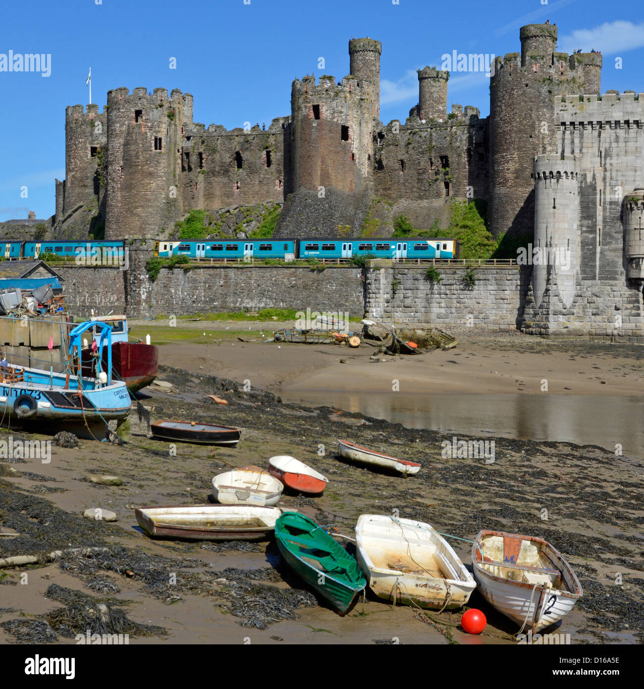 Railway line & historic Conwy Castle Arriva train exits Robert Stephenson rail bridge over River Conwy small boats on foreshore Clwyd North Wales UK Stock Photo