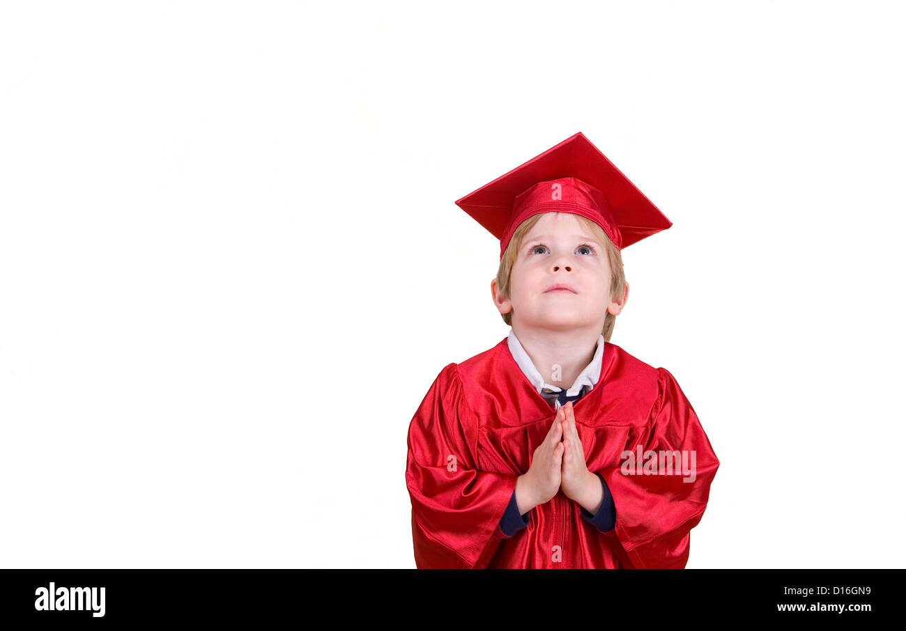 A young boy looks up with his hands in a prayer postion wearing a cap and gown. Stock Photo