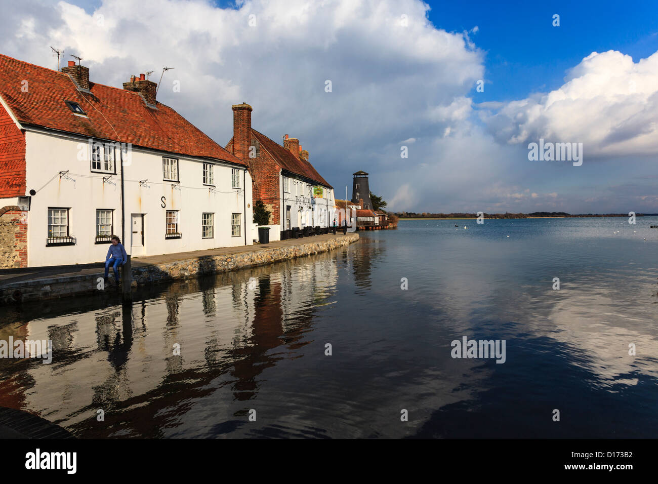 A young girl sits on a gate and swings at High Tide, Langstone, with the Royal Oak Pub and reflections in the water, Hampshire Stock Photo