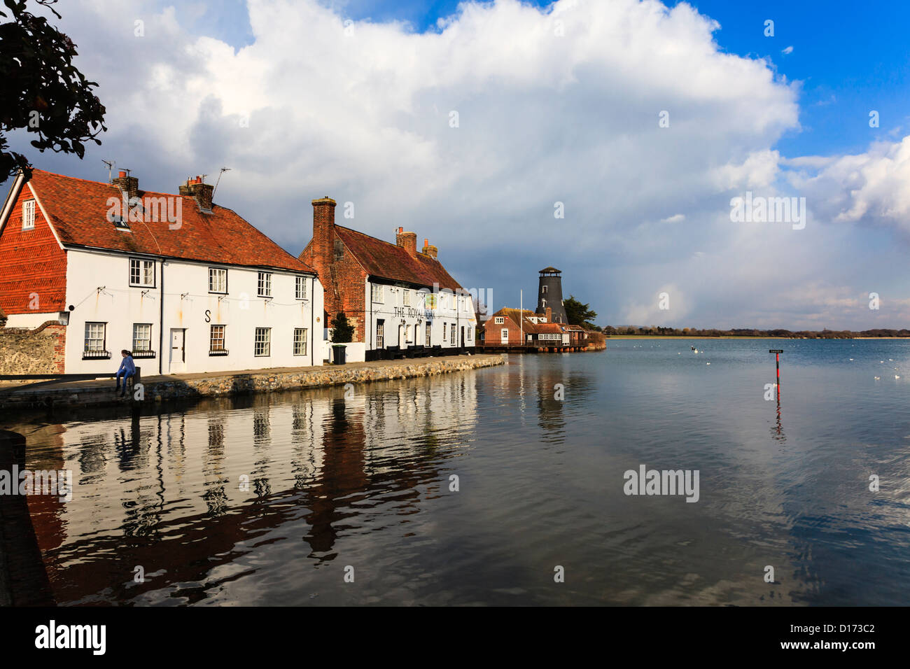 A young girl sits on a gate and swings at High Tide, Langstone, with the Royal Oak Pub and reflections in the water, Hampshire Stock Photo