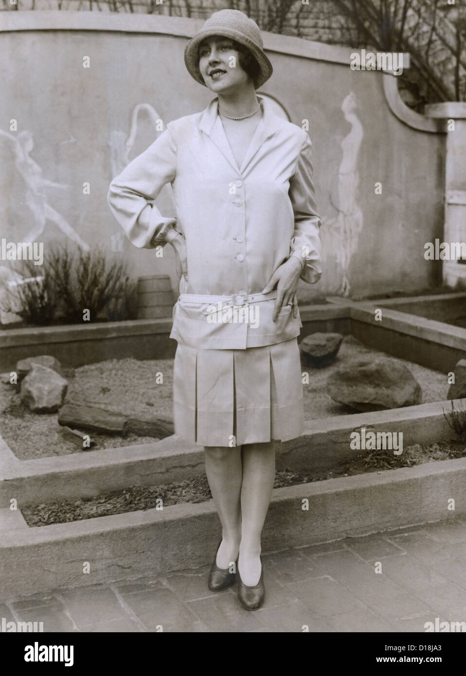 1927 suit with a mid-knee pleated skirt and matching waistless, hip-belted jacket. The outfit is toped by a cloche hat. March Stock Photo