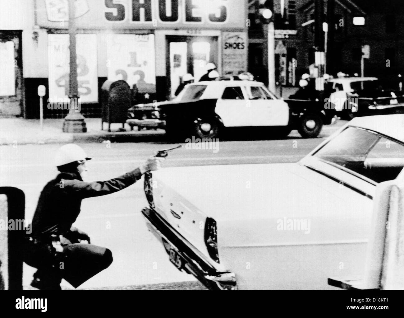 1965 Watts Riots. A policeman aims his revolver at building where a sniper shot at passing cars. Across the street, fellow Stock Photo