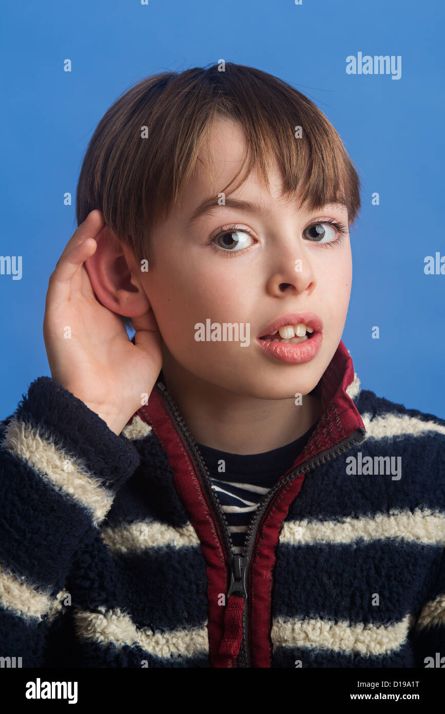 Boy aged 8 on a blue background cupping ear to improve hearing. Showing use of the sense to hear. Stock Photo