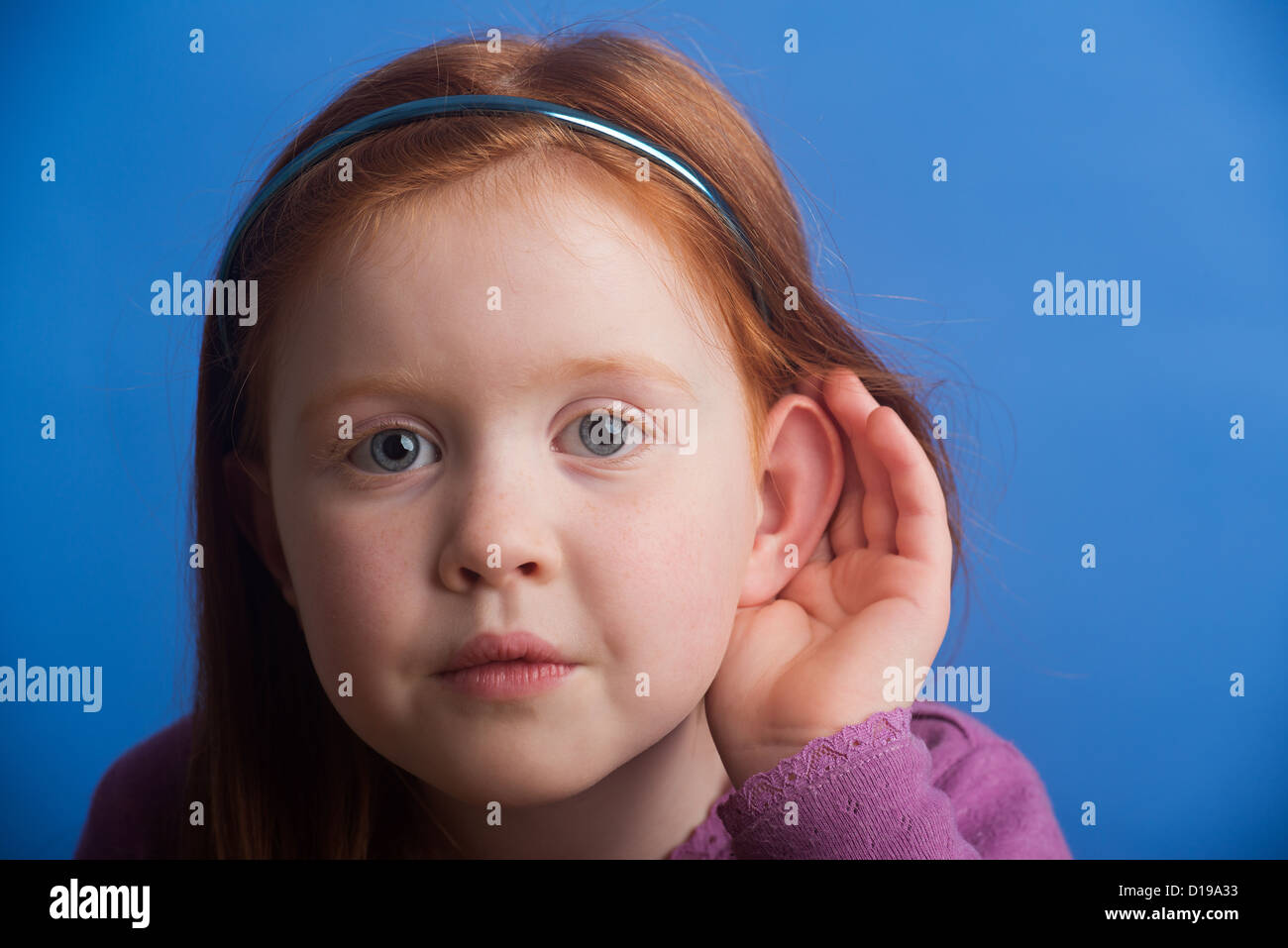 Girl aged 6 on a blue background cupping ear to improve hearing. Showing use of the sense to hear. Stock Photo