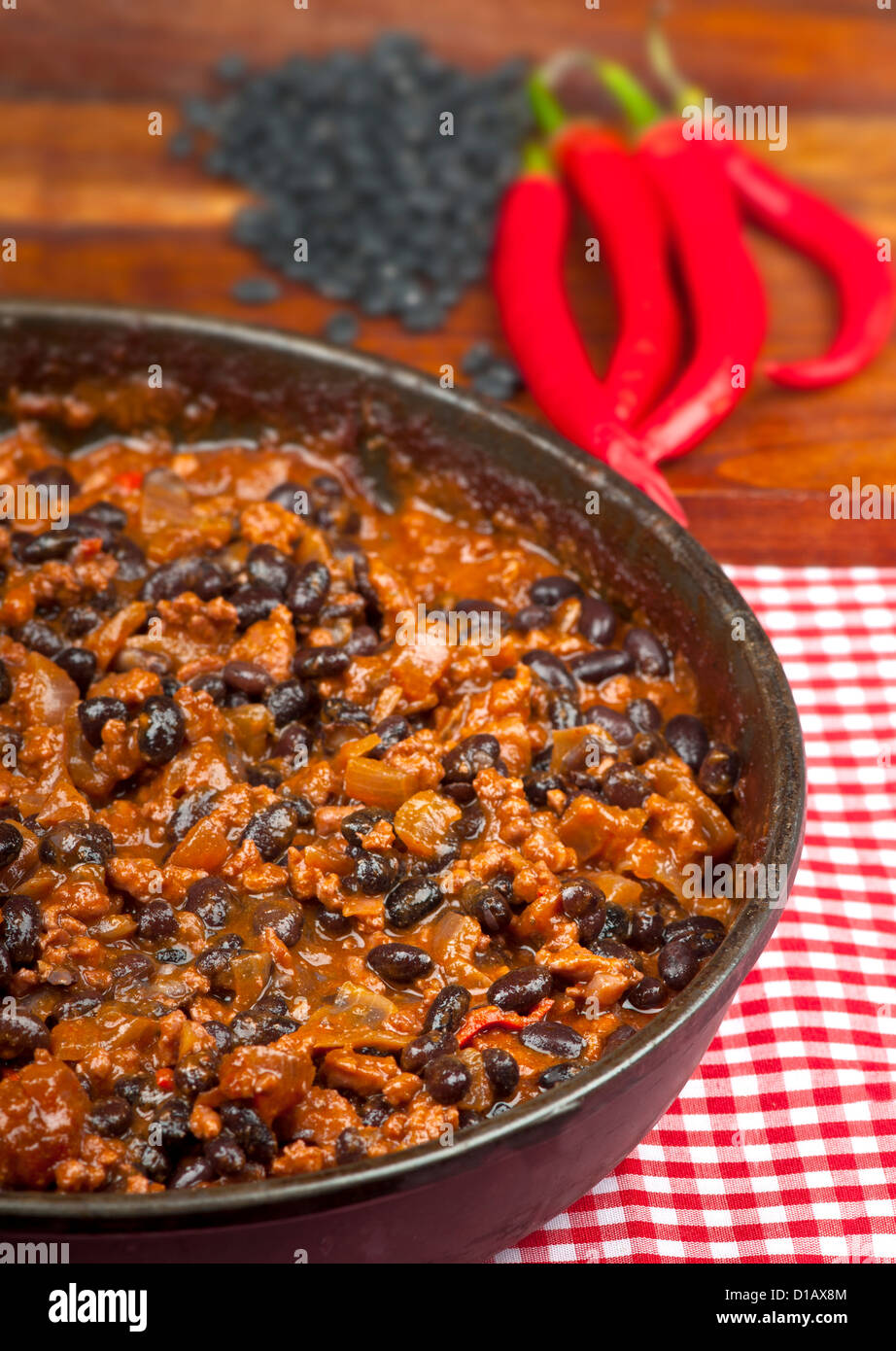 Hot and spicy Chili con Carne in the frying pan with rice. Whole red peppers and raw black beans in the background. Stock Photo
