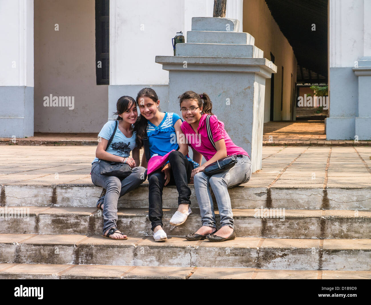 Three high school girlfriends sit together, facing forward and smiling on the steps of the Catholic church in Capiatá, Paraguay. Stock Photo