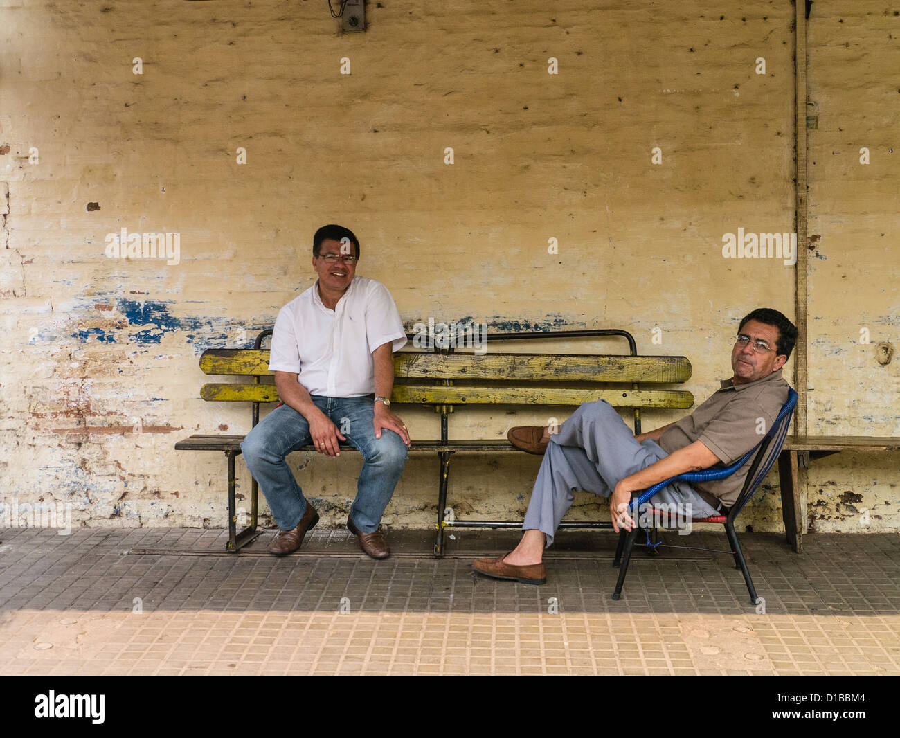 Two Hispanic men sit and talk as they take a break in the shade by a wall in Capiatá, Paraguay. Stock Photo