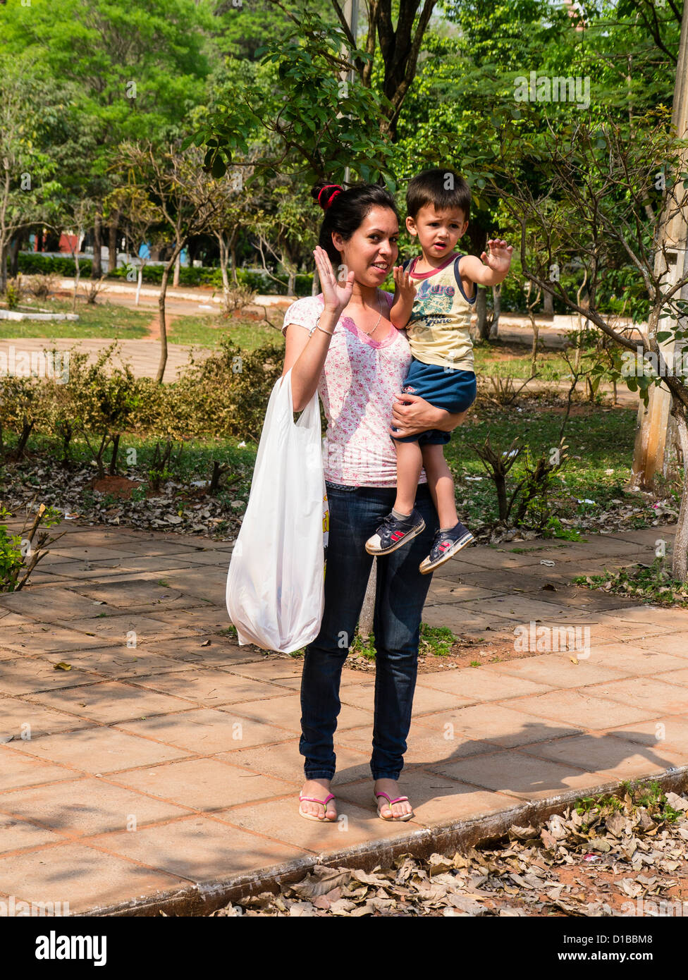 A young Hispanic mother holds her young son in one arm, while carrying a shopping bag on the other arm waving and smiling. Stock Photo