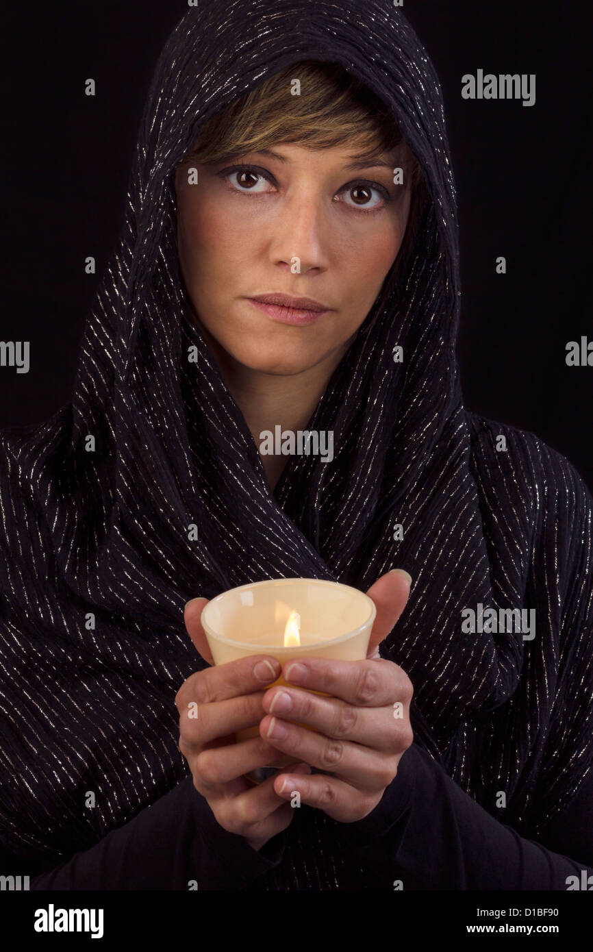 Beautiful young woman in black cape holding a burning candle in her hands and looking thoughtful at the camera. Studio portrait Stock Photo