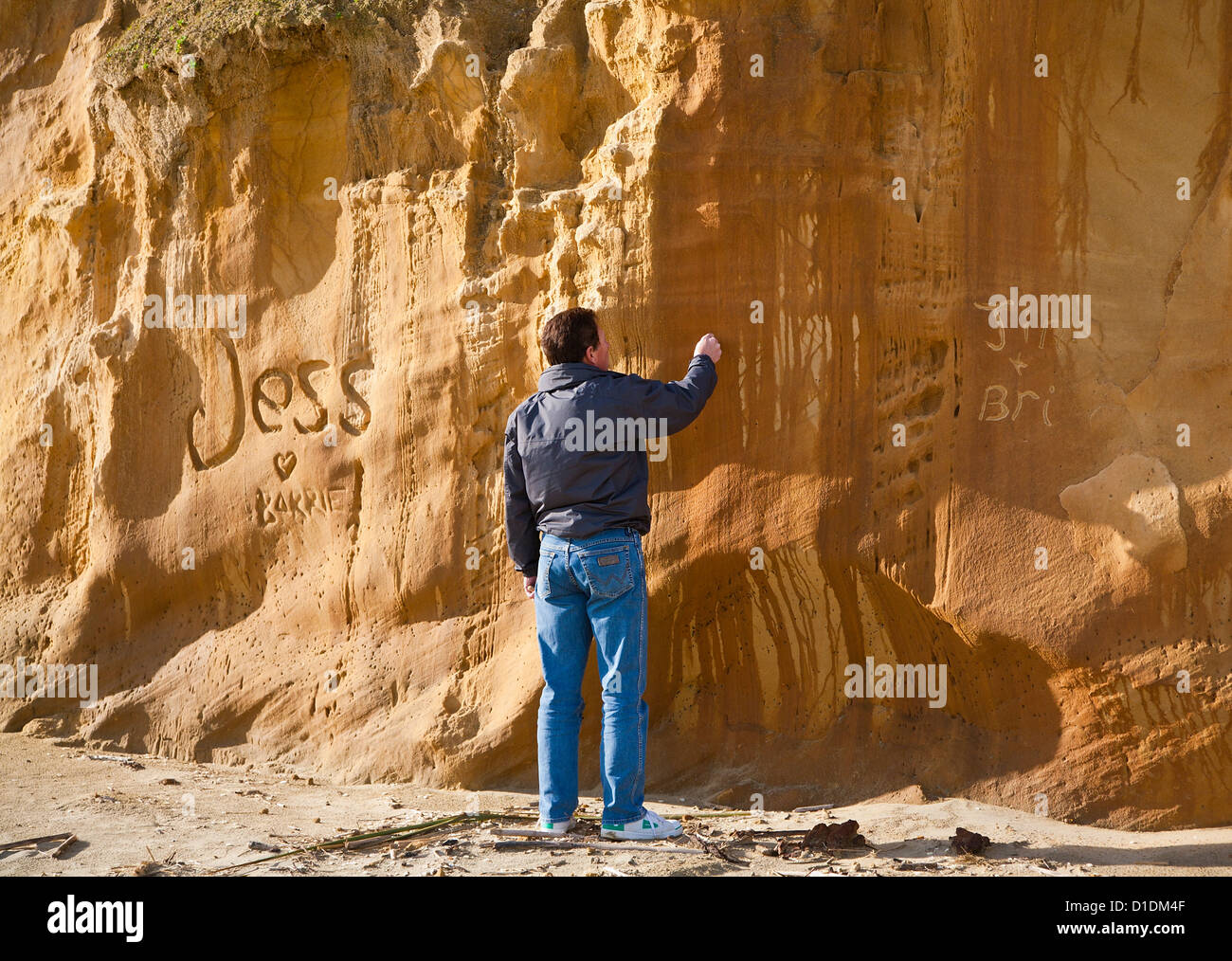 A man carving graffiti into the sandstone rocks, cliffs, at Baylys Beach, Northland, North Island, New Zealand Stock Photo