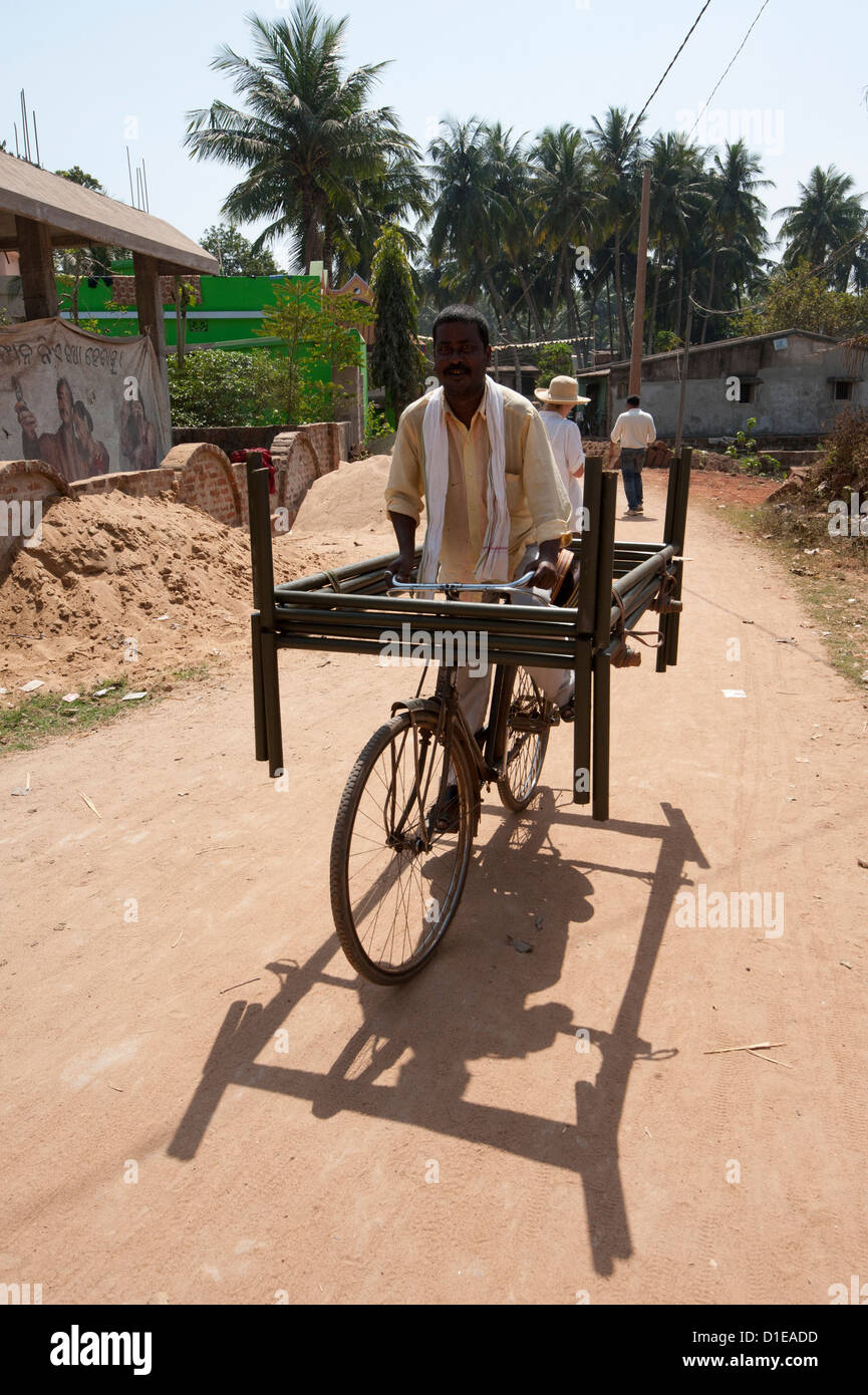 Man cycling through village street with two metal bed frames balanced on his bicycle, Hirapur, Orissa, India, Asia Stock Photo