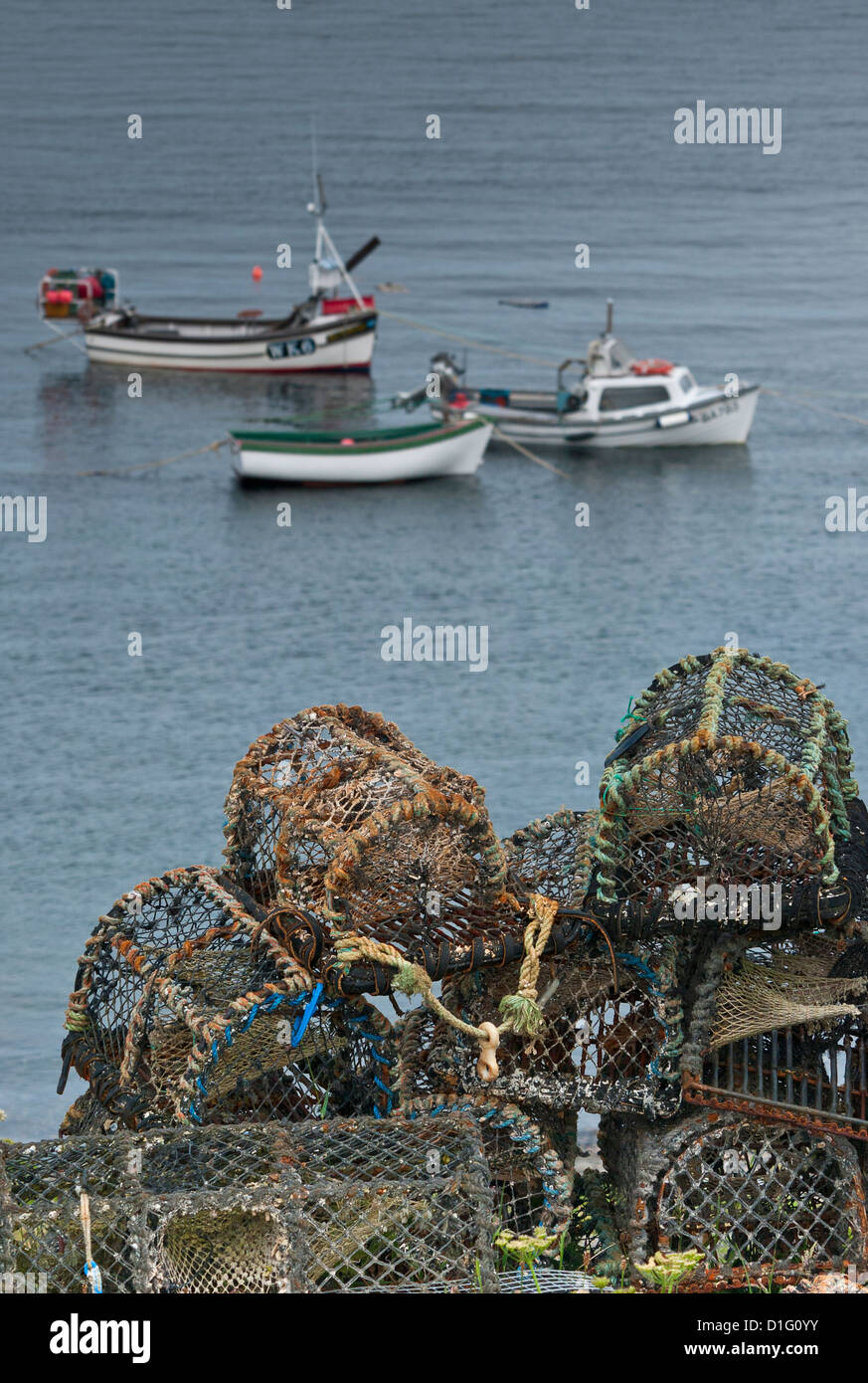 Lobster pots stacked on land, with three small fishing boats at anchor in harbour in background. Stock Photo