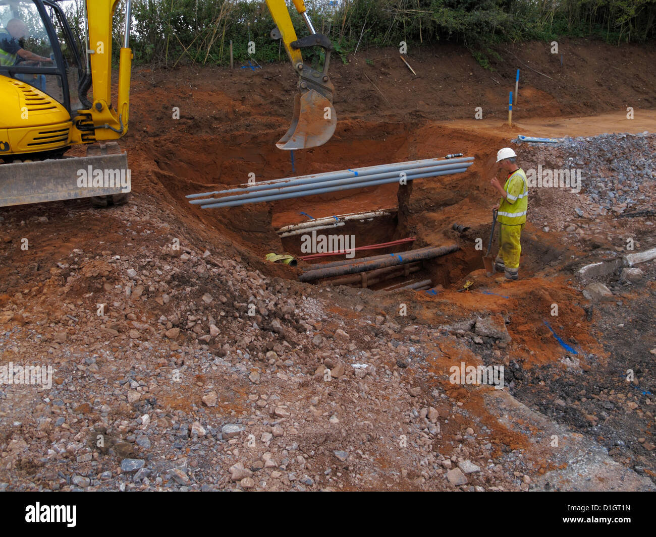 Excavator digging a trench watched by banksman underground services with a tight packed group of grey fiber-optic cable ducts uk Stock Photo