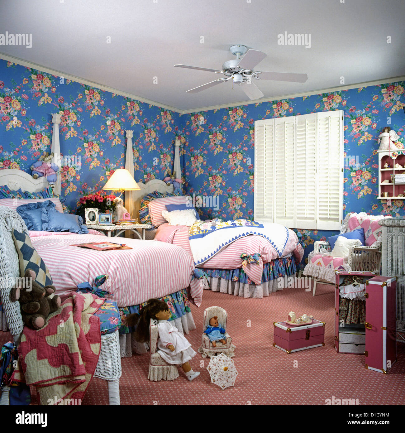 CHILDREN'S BEDROOM: Girl's twin two poster beds, blue and pink floral walls. Pink rug. White wood shutters at window. Dolls. Stock Photo