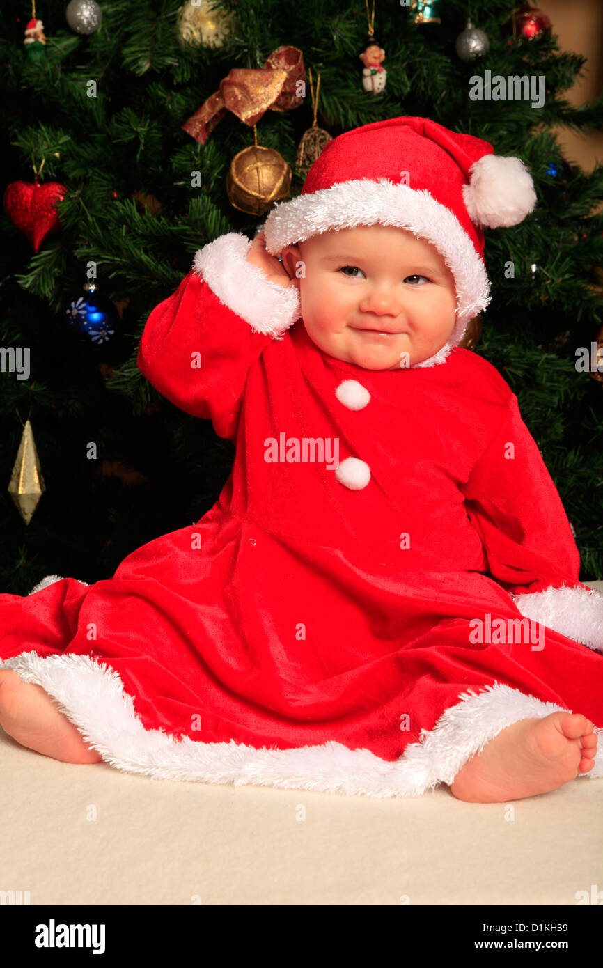 8 month old baby girl dressed like Santa Claus in front of a Christmas tree Stock Photo