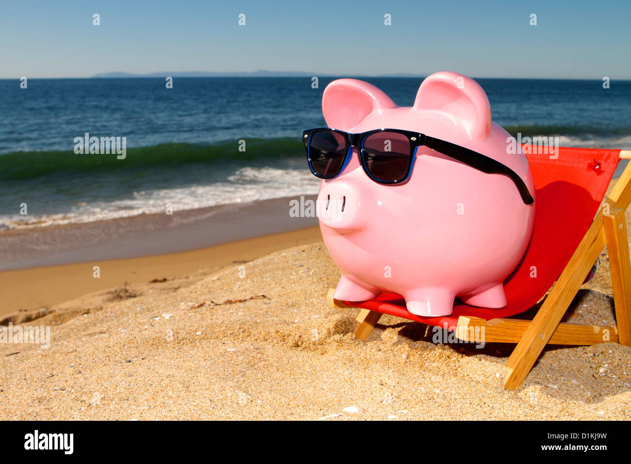 Pink piggy bank on a beach in a deck chair wearing sunglasses with golden sand a blue ocean and vivid blue sky Stock Photo