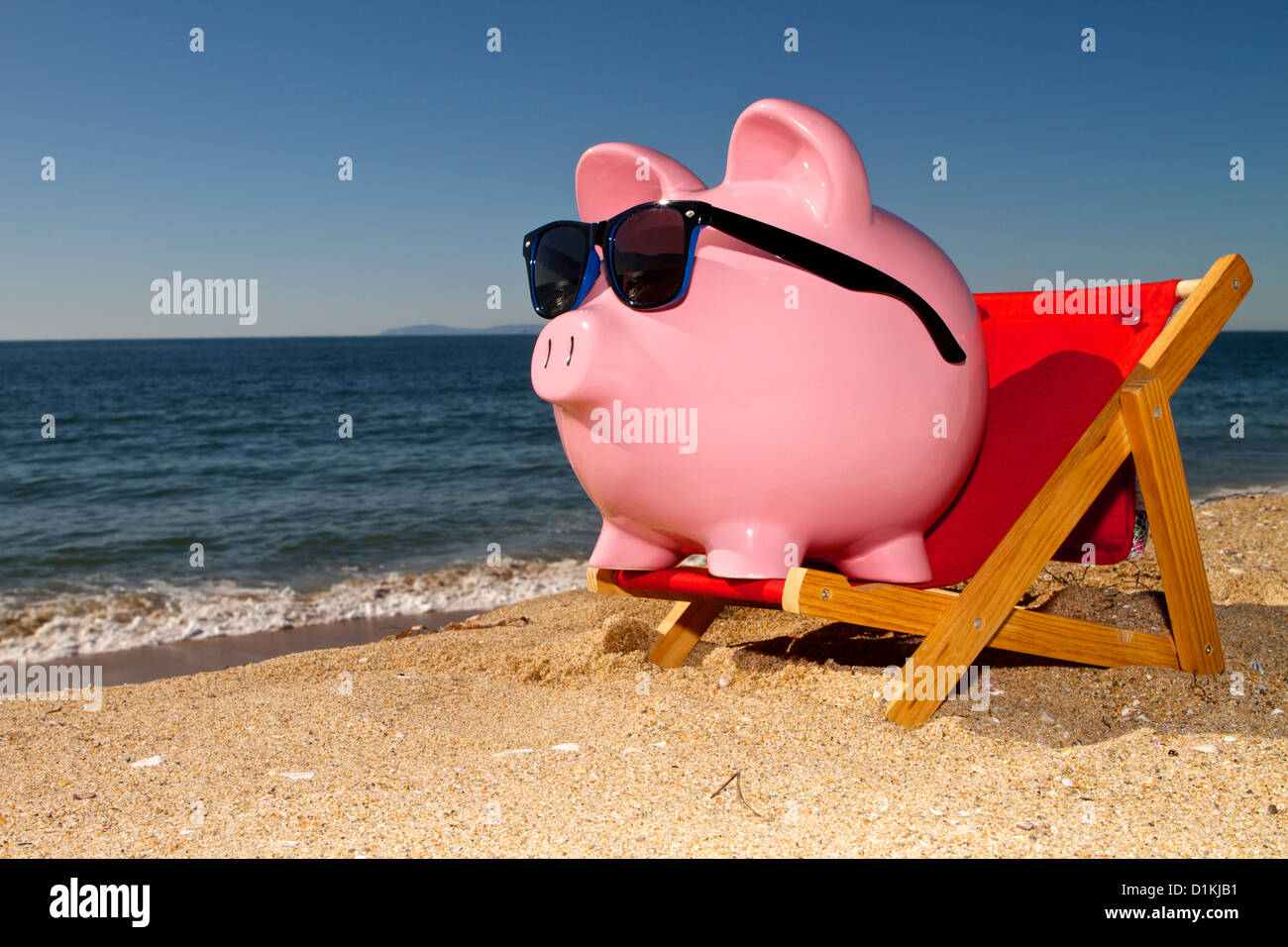 Pink piggy bank on a beach in a deck chair wearing sunglasses with golden sand a blue ocean and vivid blue sky Stock Photo