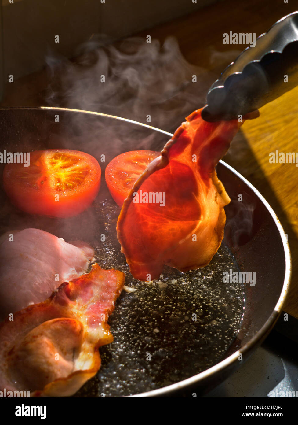BACON RASHER FRYING PAN Shaft of sunlight illuminates a rasher of organic back bacon being turned in a hot frying pan containing tomatoes Stock Photo
