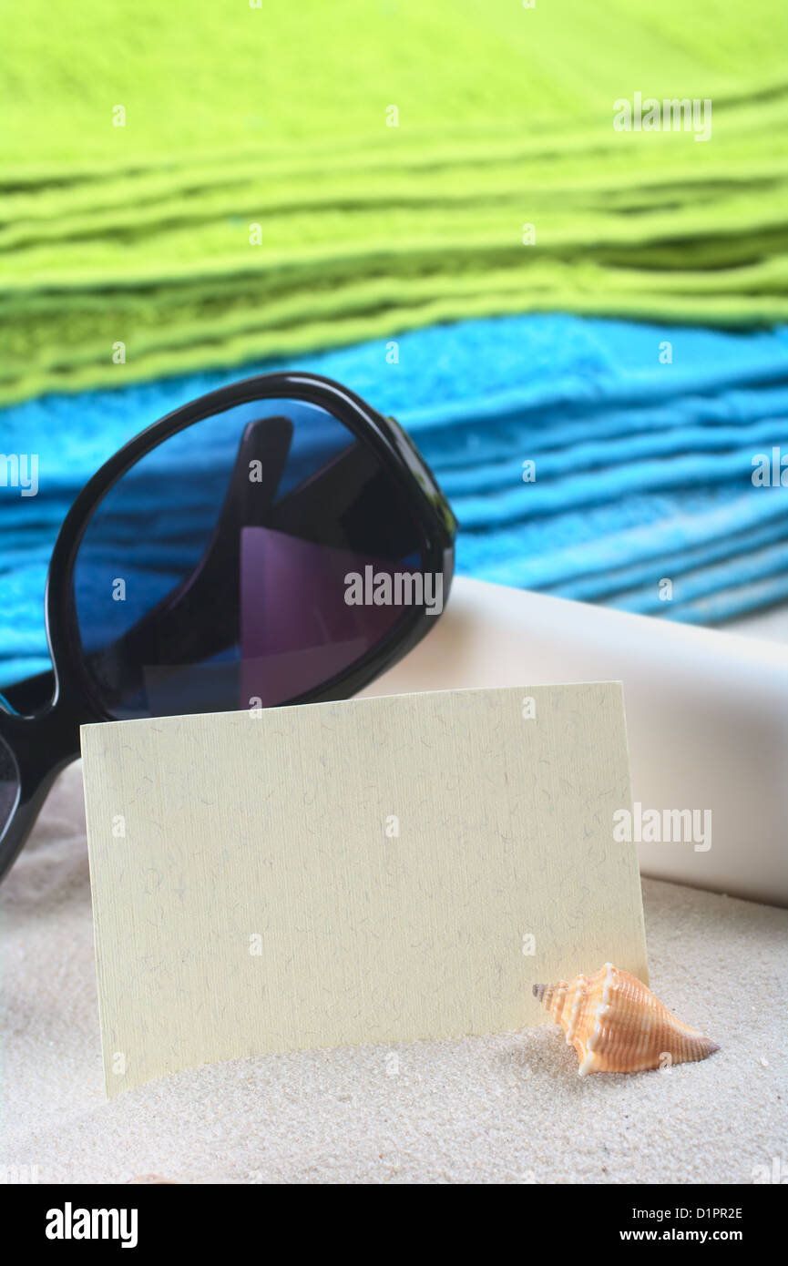 Blank card on sand with a shell in front, and sunglasses, suncream and colorful beach towels in the back Stock Photo