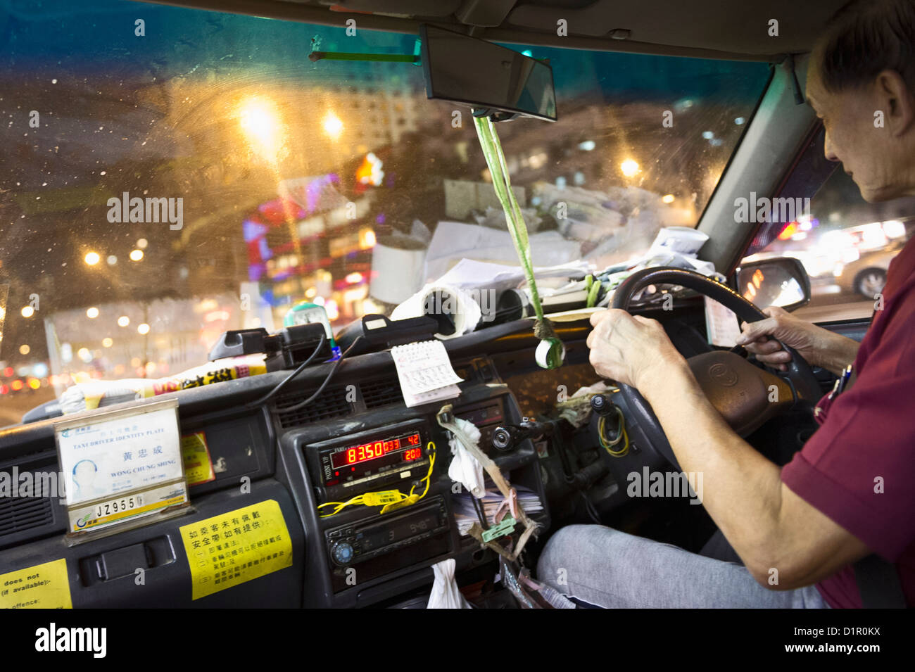 A Hong Kong taxi driver is seated behind the wheel, with dashboard-mounted mobile devices visible, as city traffic and colorful lights illuminate the night outside.