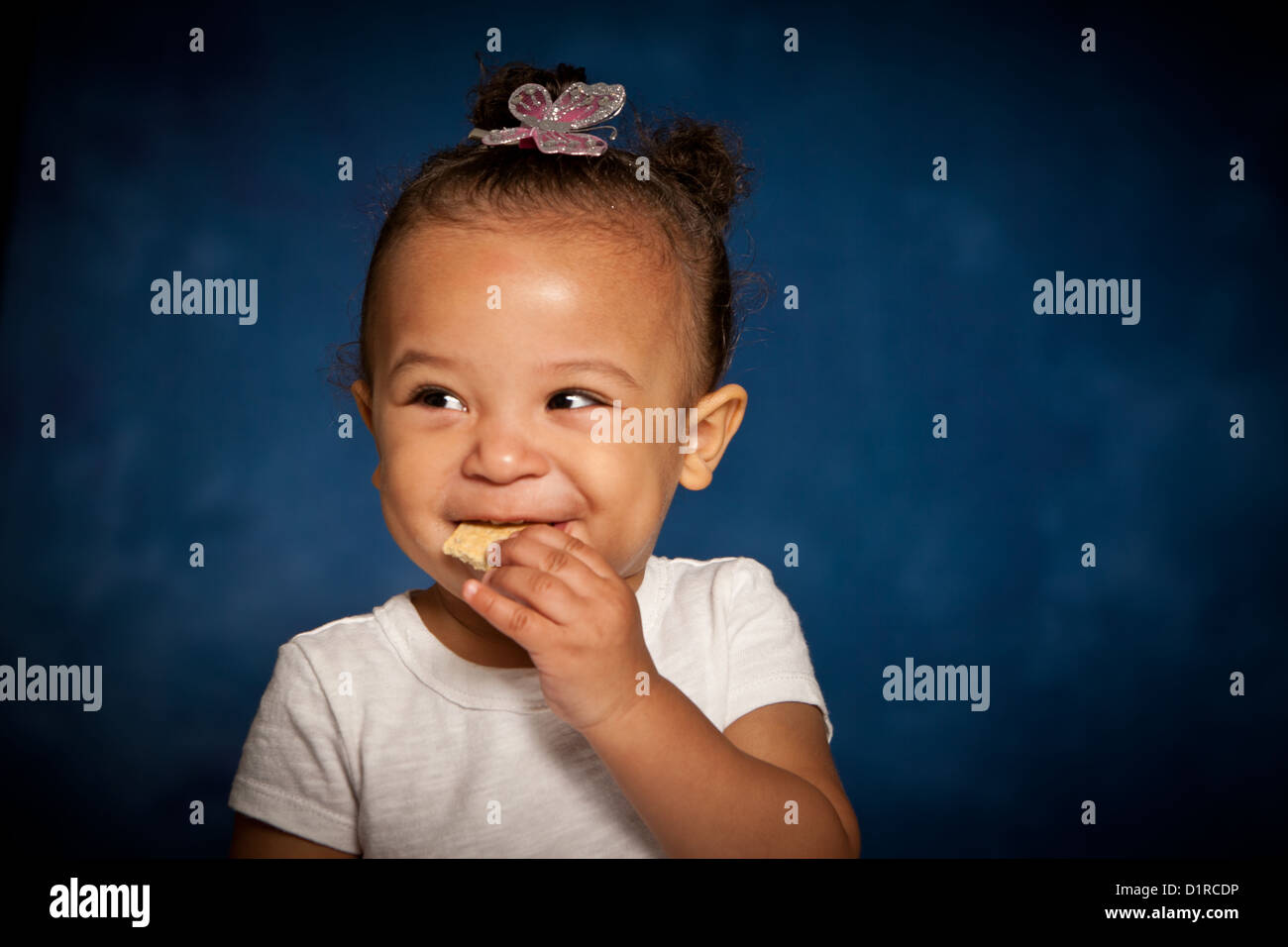 Cute studio portrait of a mixed race toddler girl eating a biscuit with a cheeky smile on her face Stock Photo