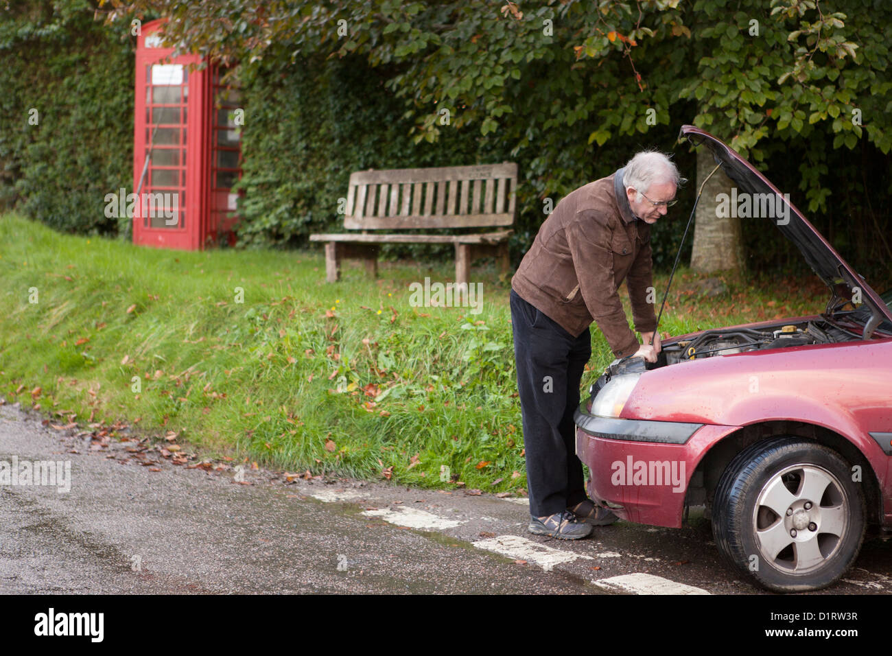 Man looking under bonnet of broken down car at side of road with telephone box and bench in background Stock Photo