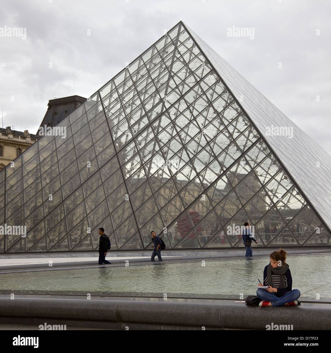 A young woman sits cross legged and writes in a notepad in front of the Louvre glass pyramid Paris, Ile de la Cite, France Stock Photo