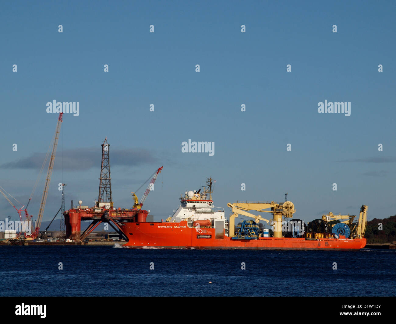 A North Sea construction vessel pass the Nigg Energy Park, with an oil rig in the dry dock, on route to Invergordon Stock Photo